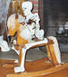 Photo of Skeleton on child's wooden rocking horse, holding skeleton-faced doll